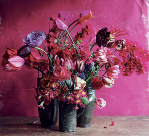 Three vases with vibrant pink, purple, and red flowers against a bright pink background on a wooden surface.