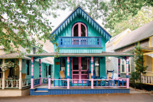 A colorful house with teal, pink, and blue trim, featuring a porch, balcony, and rocking chairs.