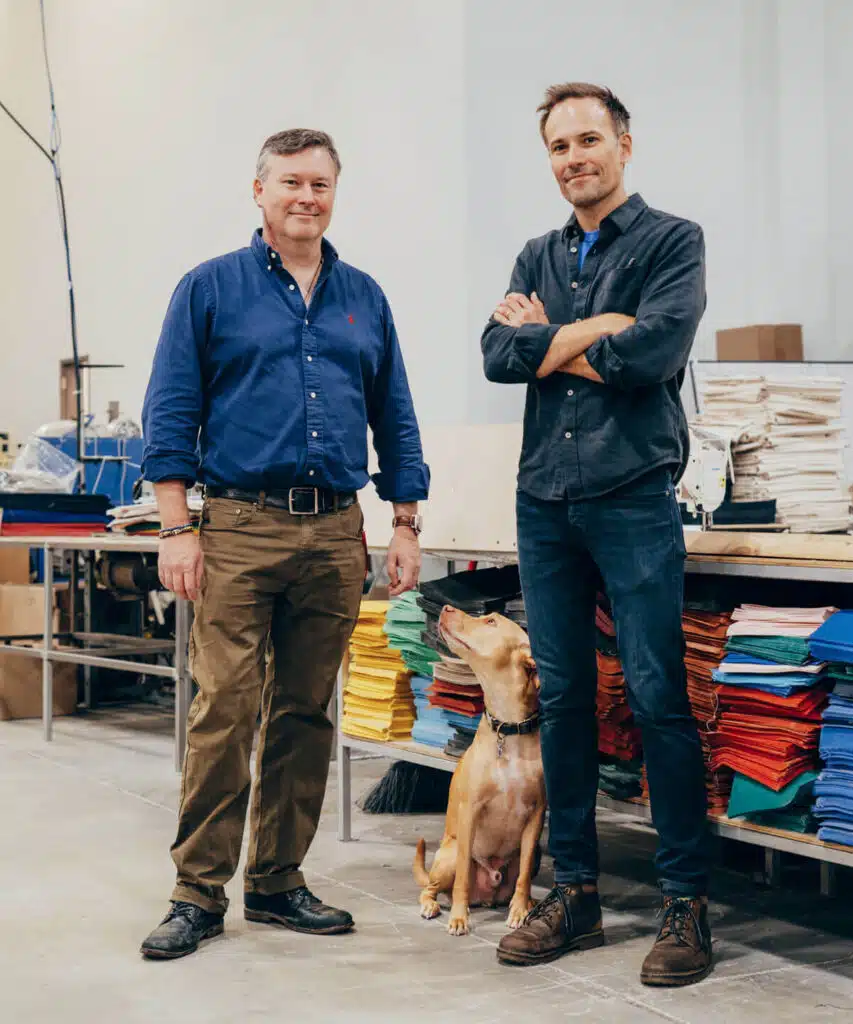 Two men stand in a workshop with colorful fabric stacks as a brown dog sits between them, looking up.