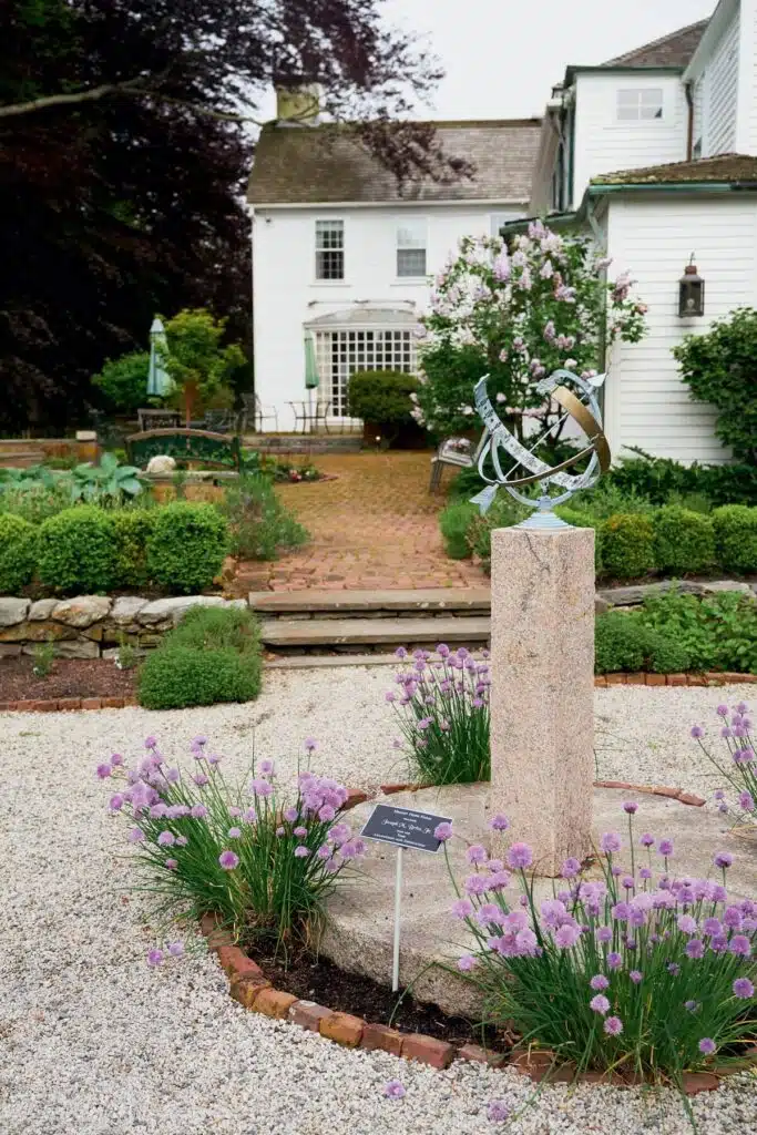New England Farm Stays. A sundial on a stone pedestal in a garden with purple flowers, in front of a white house.