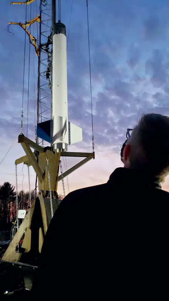 A person looks up at a rocket on a launch pad at sunset, silhouetted against a cloudy sky.