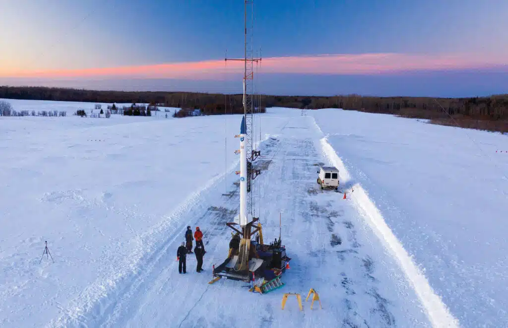 A small rocket stands on a snowy runway with people and a van nearby at sunset.