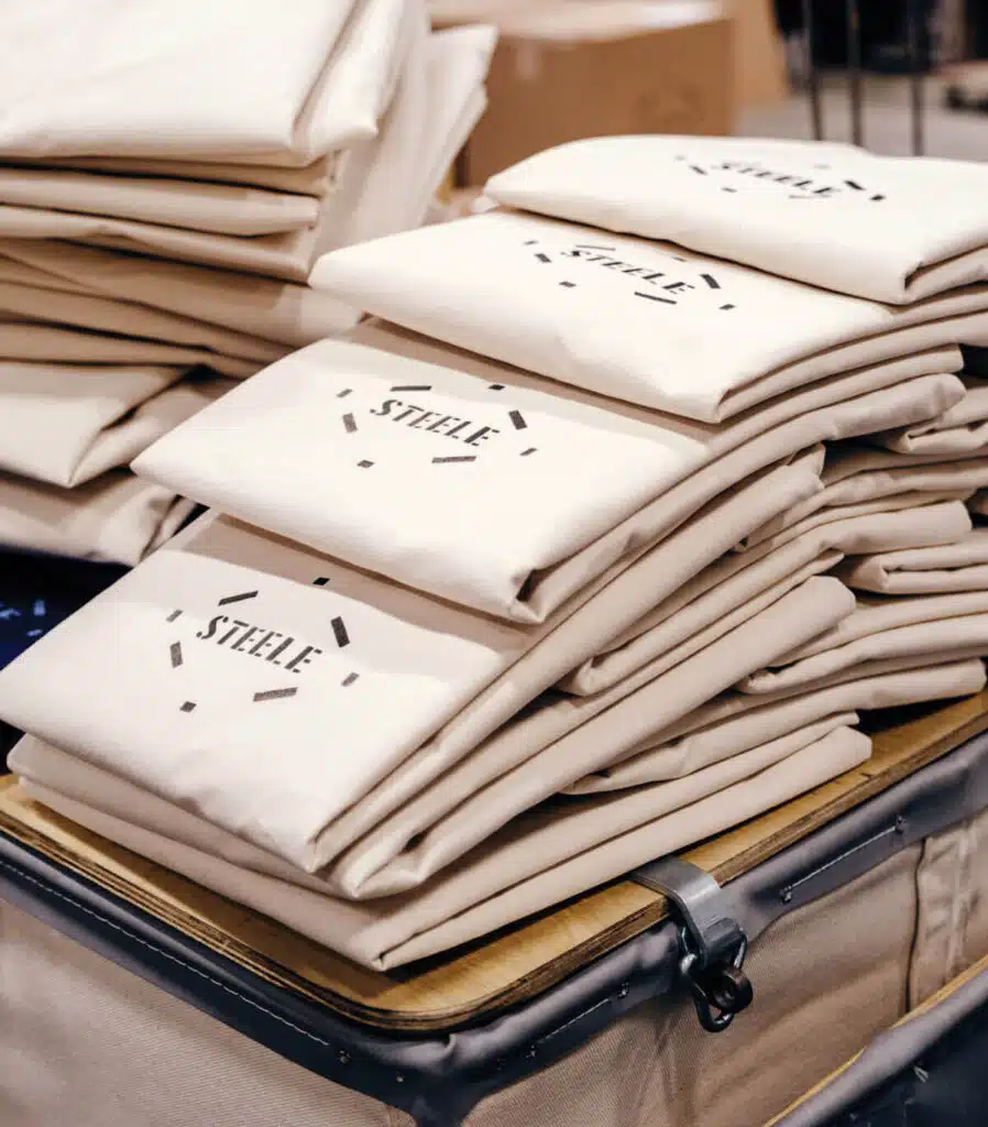 Steele Canvas Basket Corp.. Stacks of folded beige fabric bags labeled "STEELE" in a canvas truck, created by Steele Canvas.