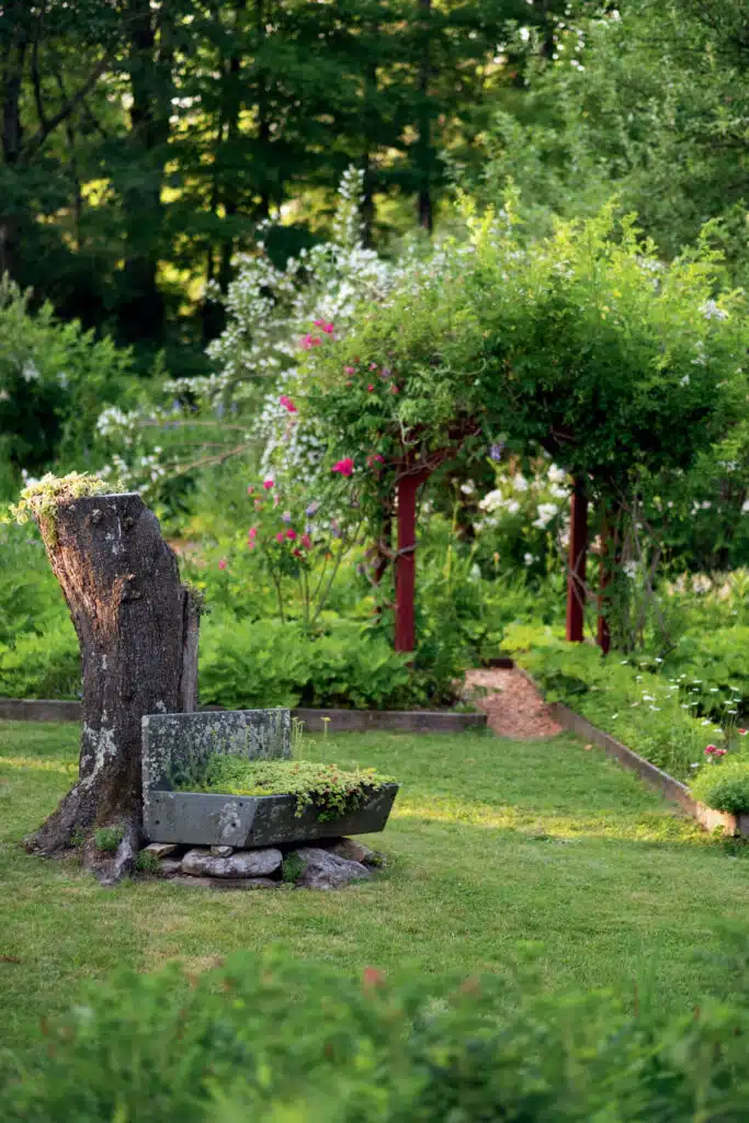 Lush garden with a tree stump planter and a flower-covered arbor surrounded by greenery.