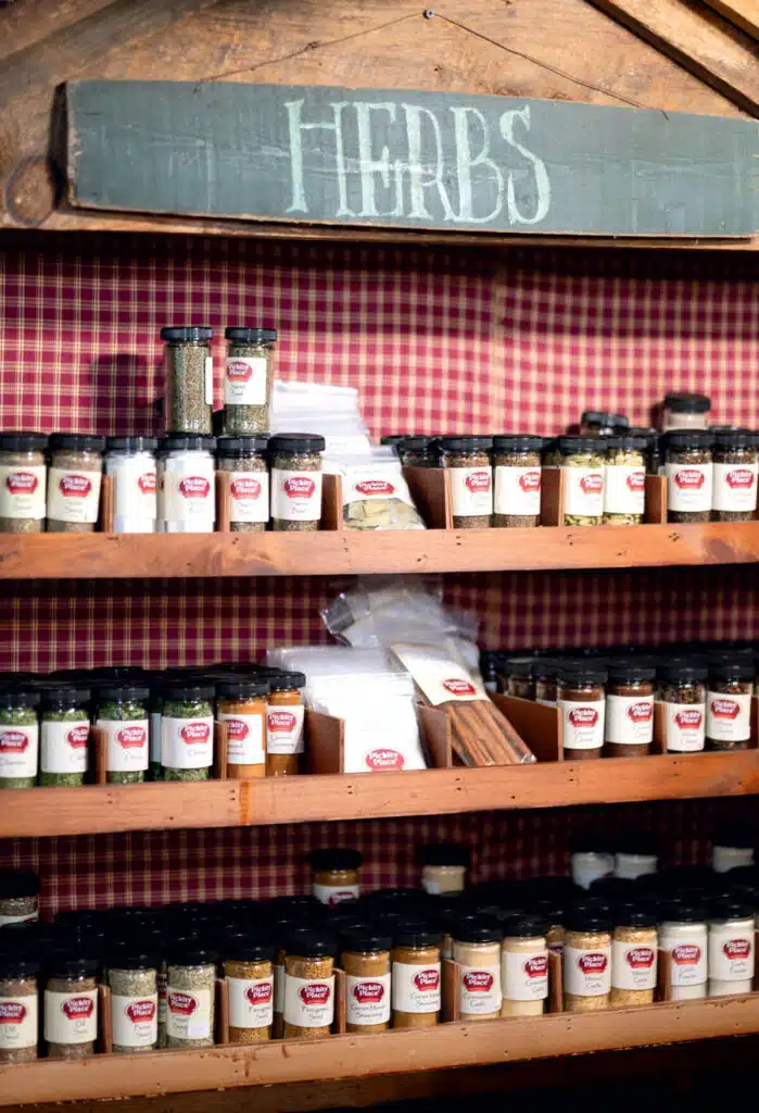 Jars of herbs and spices are neatly arranged on wooden shelves below a sign that reads "HERBS.