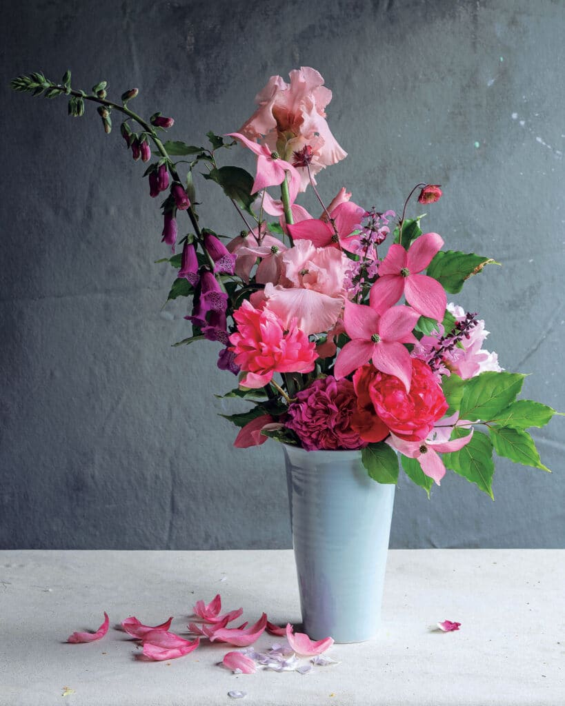 A white vase holds an arrangement of pink and purple flowers with green leaves, petals scattered below.