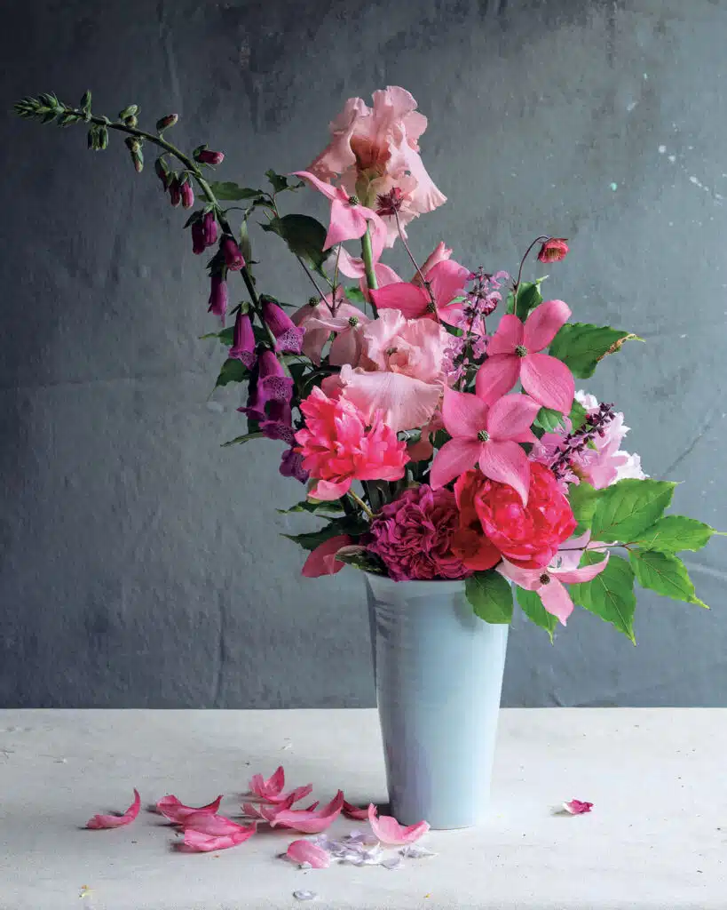 A white vase holds an arrangement of pink and purple flowers with green leaves, petals scattered below.