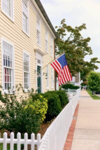 A yellow house with an American flag, white fence, bushes, and a sidewalk on a sunny day.