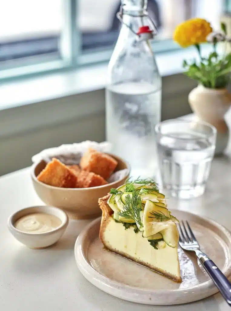 A slice of savory quiche topped with fresh dill, bread cubes, sauce, water, and flowers on a table.