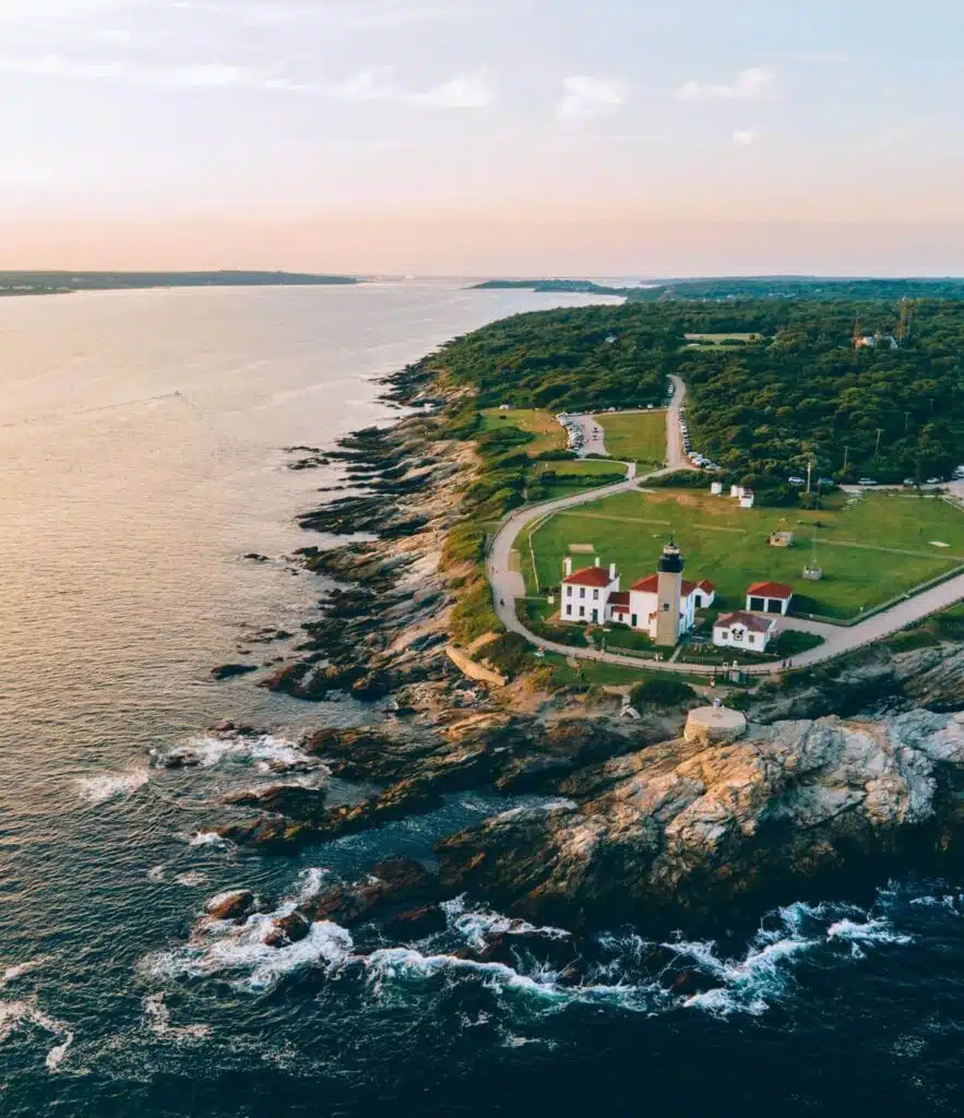 Aerial view of a lighthouse on a rocky coastline with green fields and the ocean at sunset.