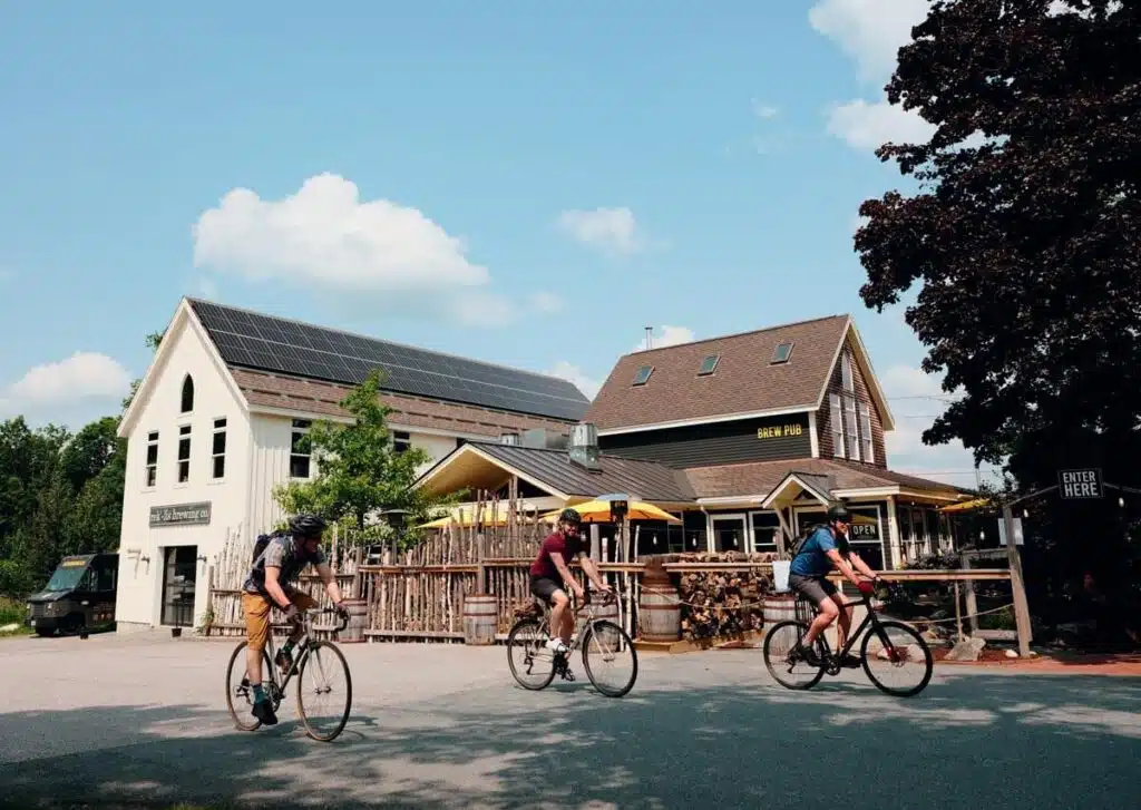 Three cyclists ride past a rustic building with outdoor seating on a sunny day.
