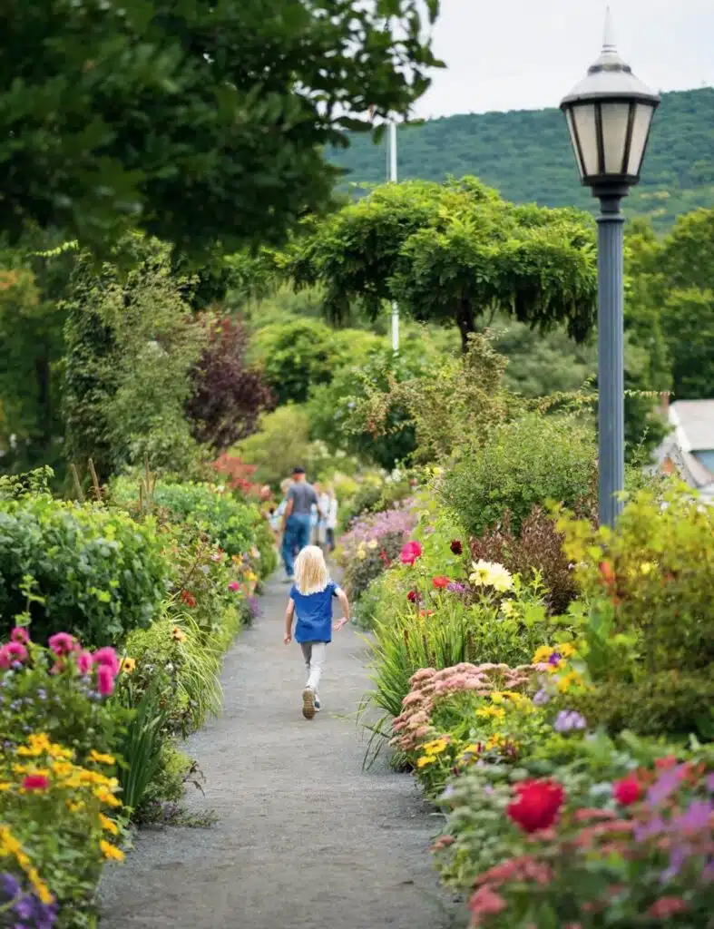 A young girl walks along a garden path lined with colorful flowers and greenery.