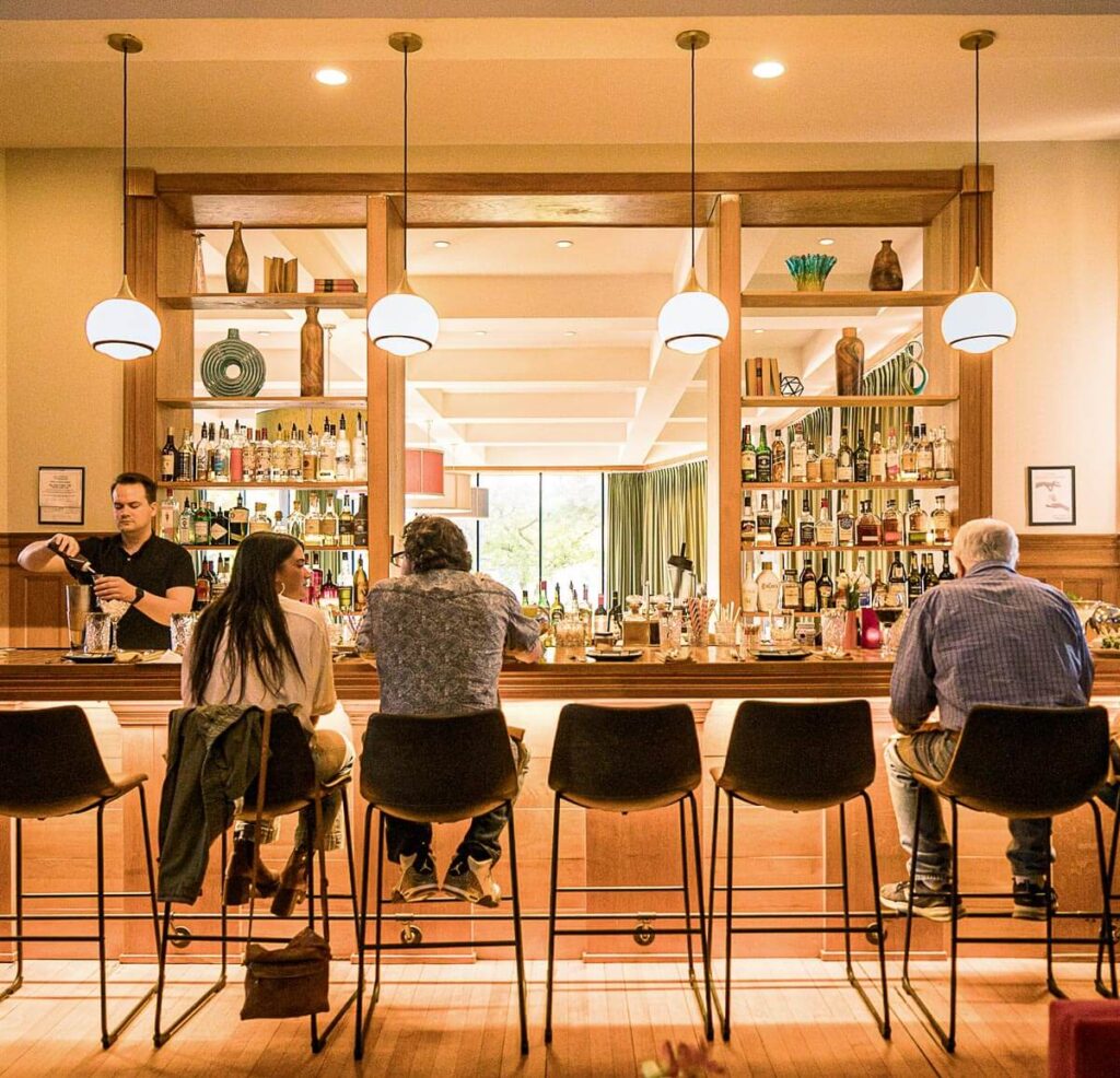Four people sit at a bar with shelves of bottles behind, while a bartender prepares a drink in warm lighting.