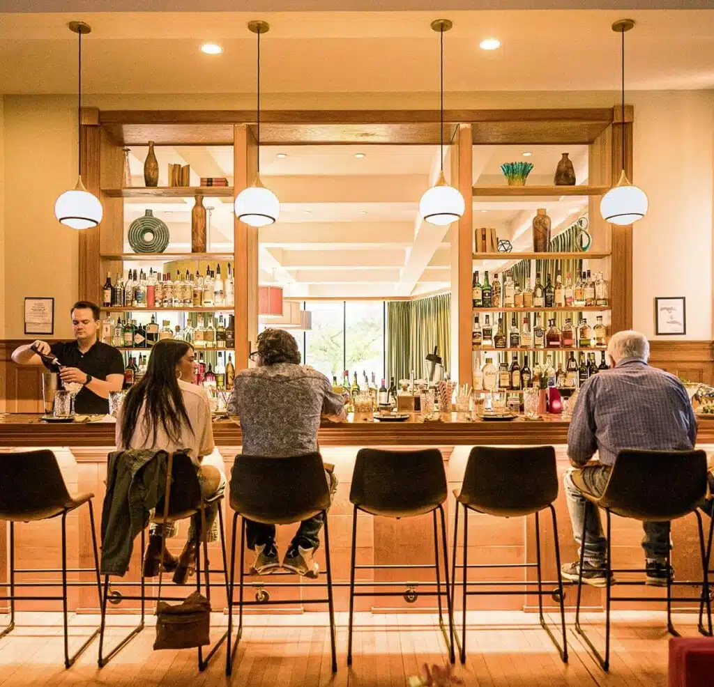 Four people sit at a bar with shelves of bottles behind, while a bartender prepares a drink in warm lighting.