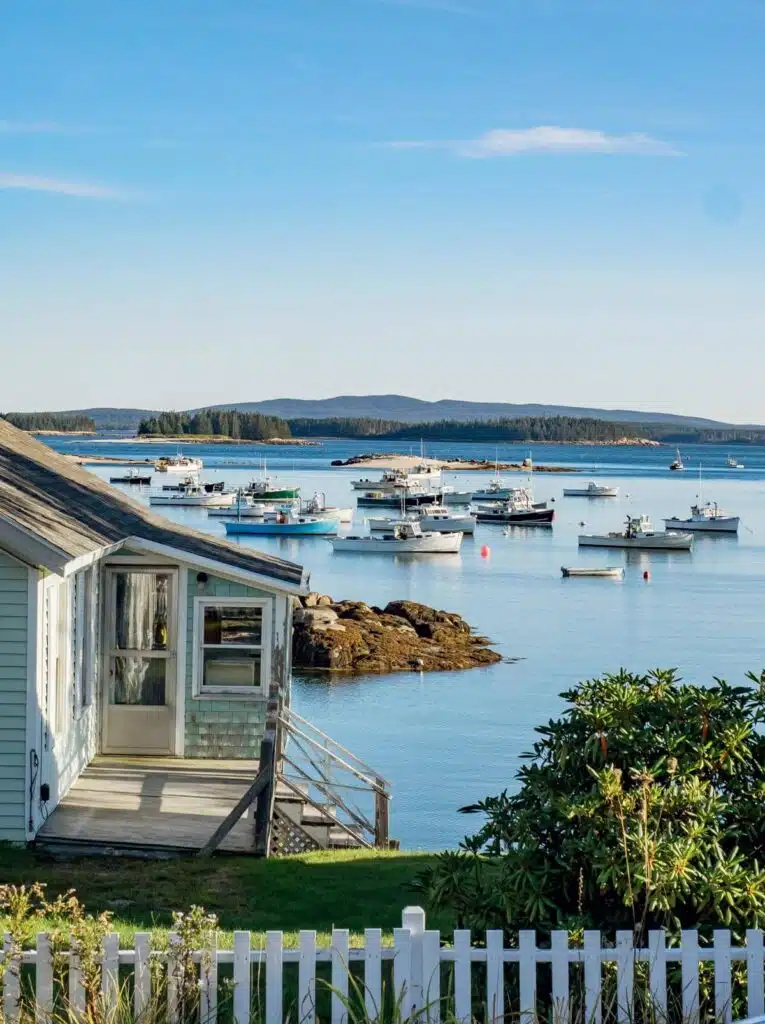 The 25 Best Small Towns in New England. Boats anchored in a calm bay near a coastal house with white picket fence, under a clear blue sky.