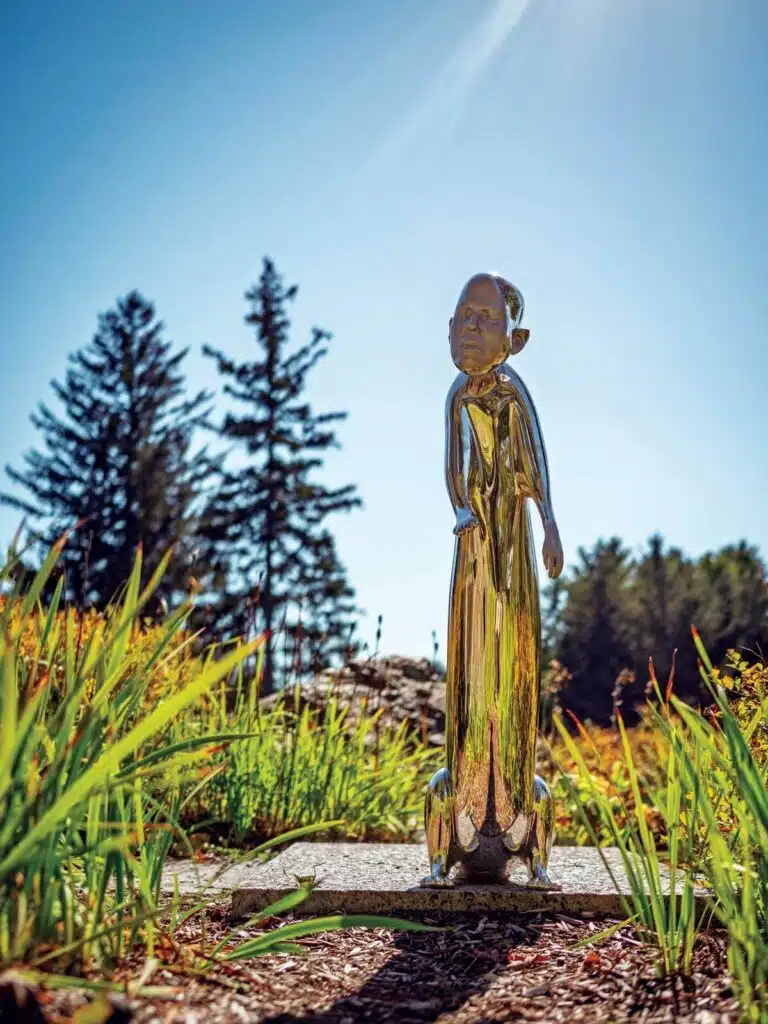 Shiny metallic human-like sculpture standing on a stone path amid green plants and trees under a clear sky.