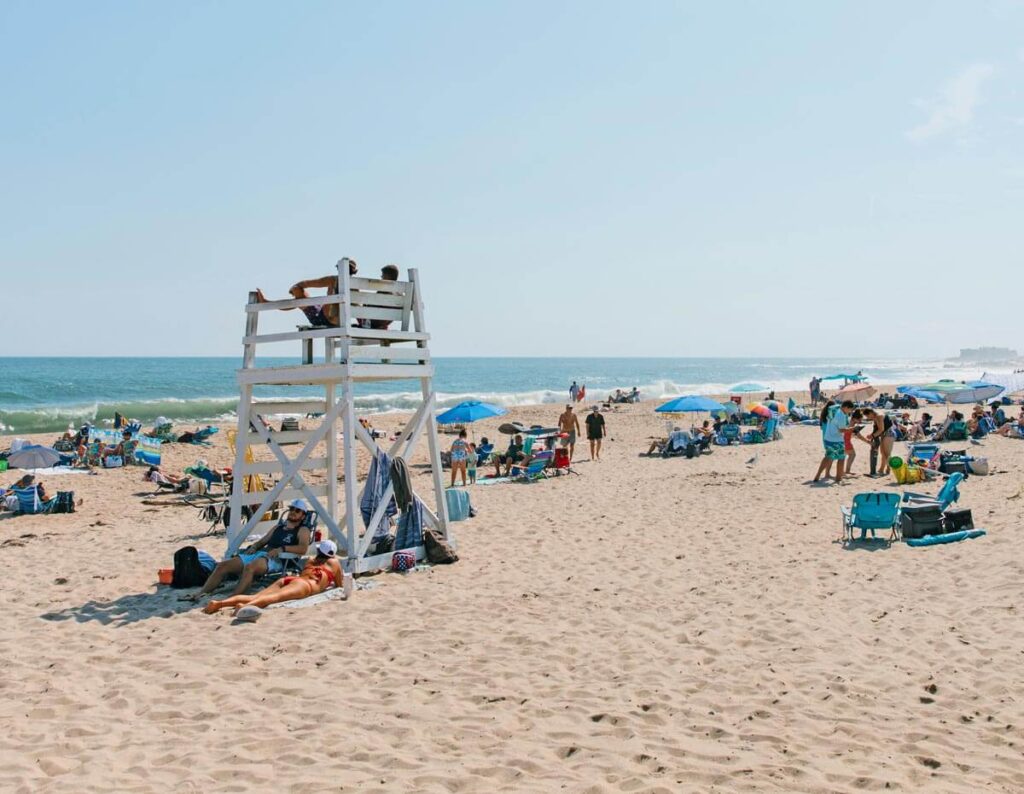 Crowded beach with people relaxing, lifeguards on a tall white chair, and umbrellas by the ocean.