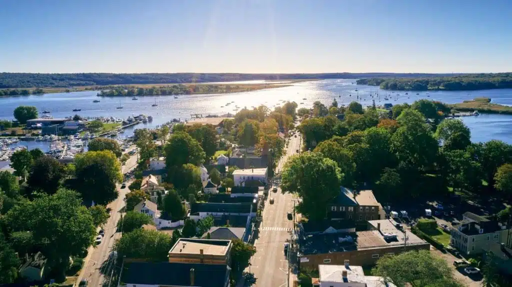 Aerial view of a riverside town with green trees, houses, and boats on a sunny day.