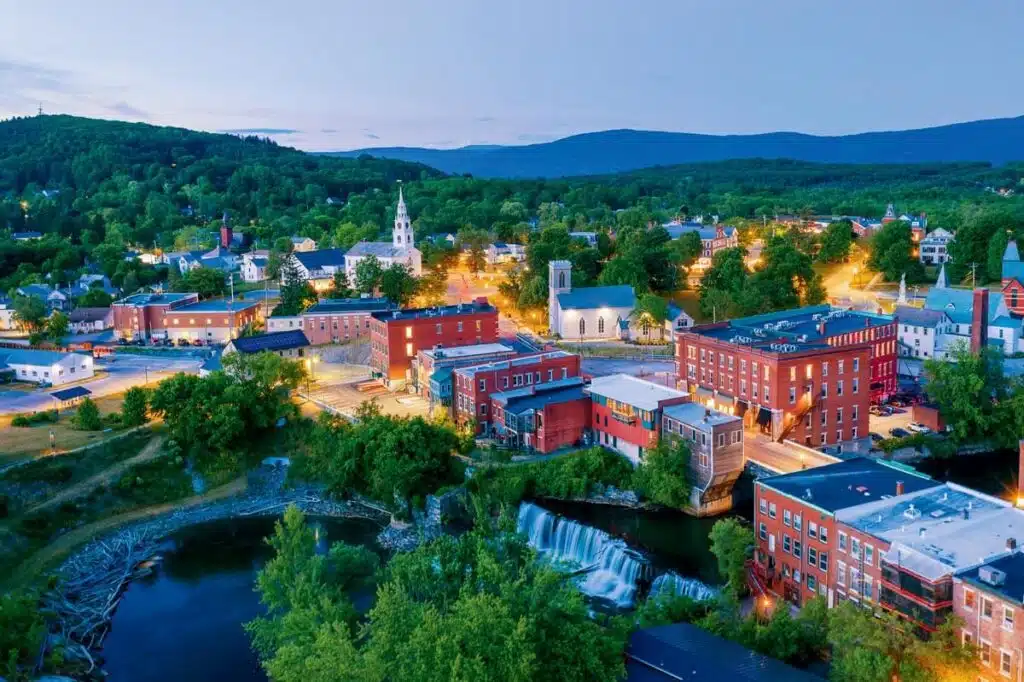 The 25 Best Small Towns in New England. Aerial view of a small town with red brick buildings, church steeple, and a river with a waterfall at dusk.