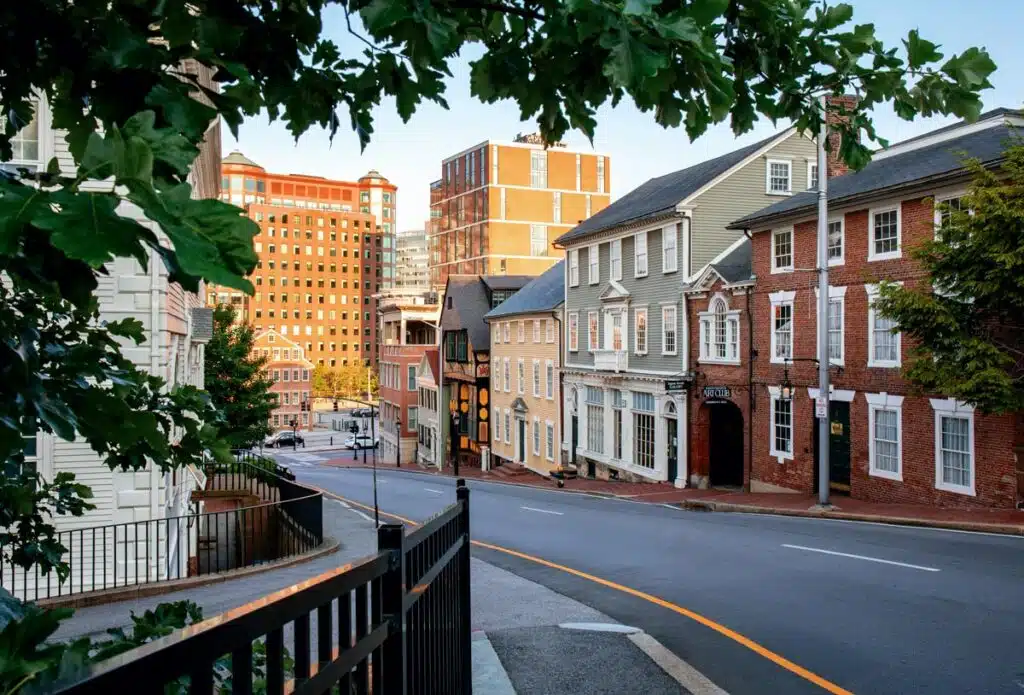 Spring Weekend in Providence, Rhode Island. Curved city street lined with historic brick and wooden buildings, framed by leafy green tree branches.