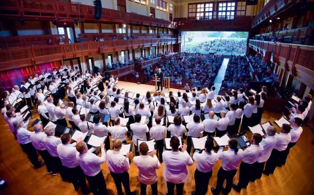 A large choir in white shirts performs on stage in a semicircle before an audience in a concert hall.