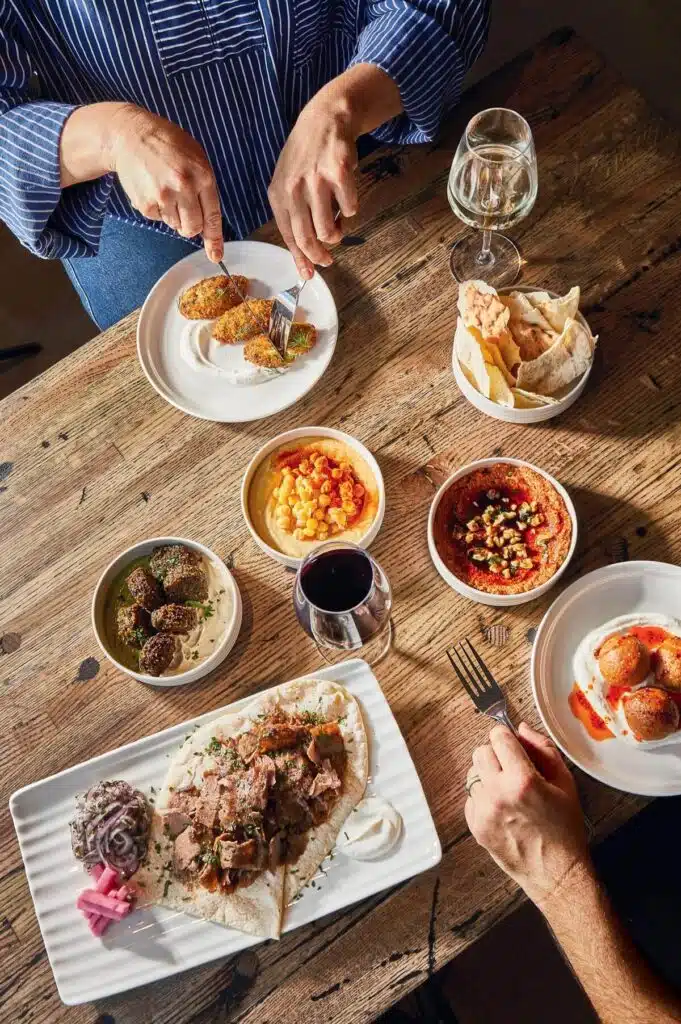 Overhead view of two people dining with various Mediterranean dishes, including pita, dips, and wine on a wooden table.