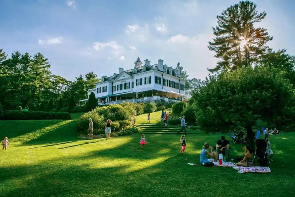The 25 Best Small Towns in New England. People relaxing on a grassy lawn in front of a large white mansion surrounded by trees on a sunny day.