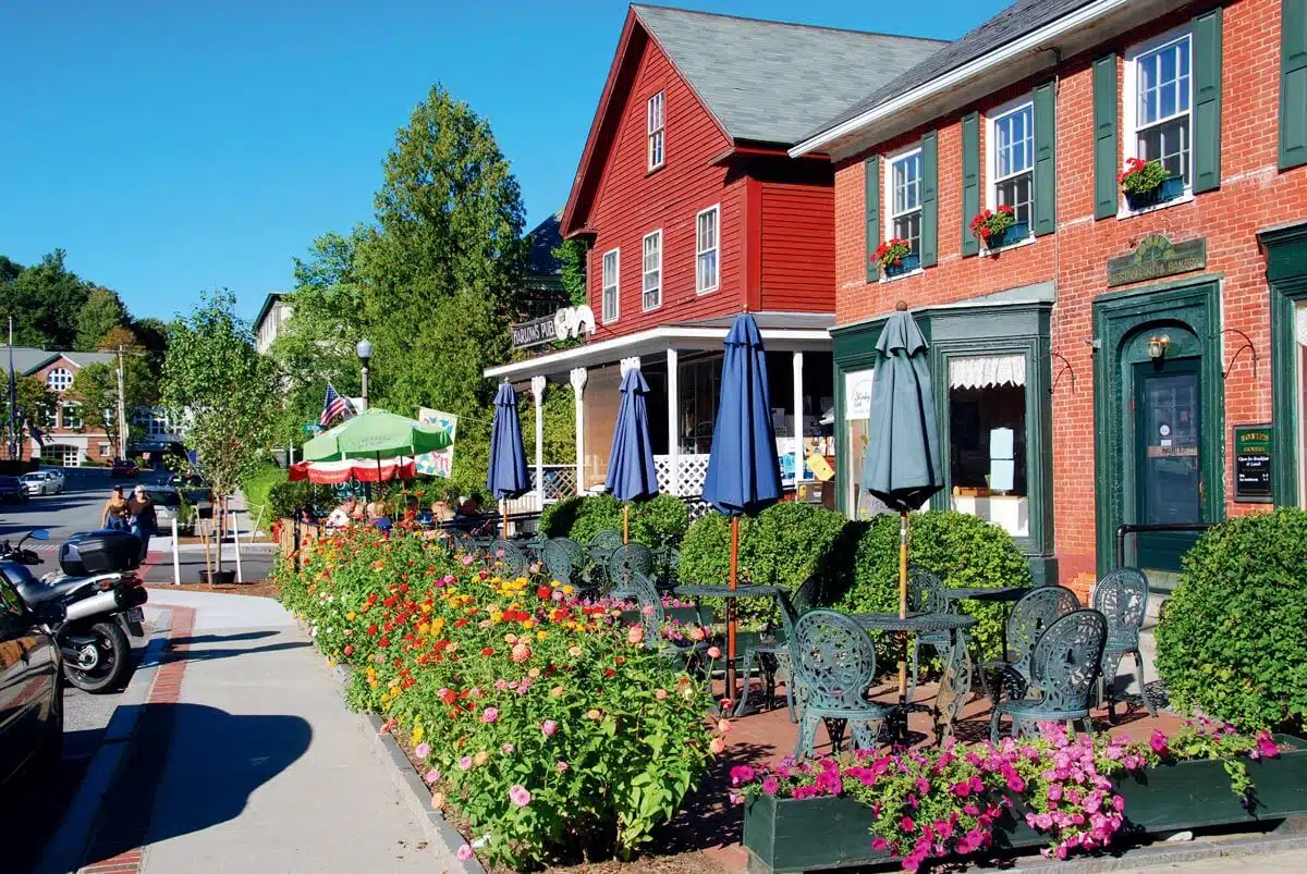 Outdoor café tables with umbrellas along a flower-lined sidewalk in a charming small town under a blue sky.