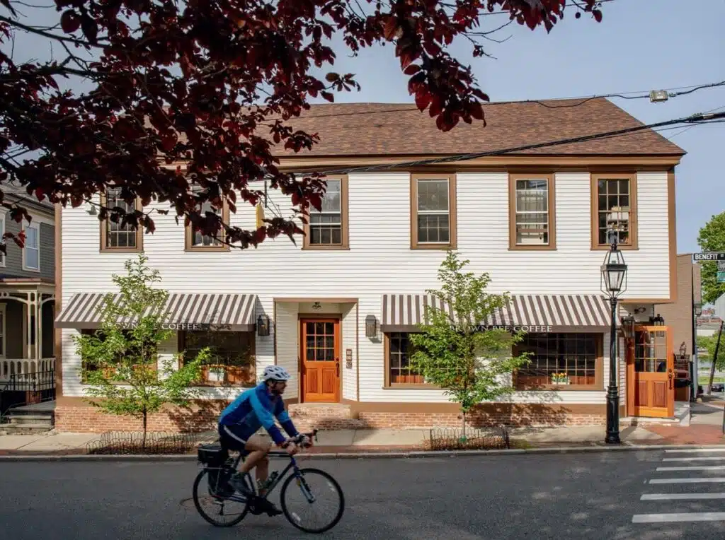 A person rides a bicycle past a coffee shop with striped awnings on a tree-lined street corner.