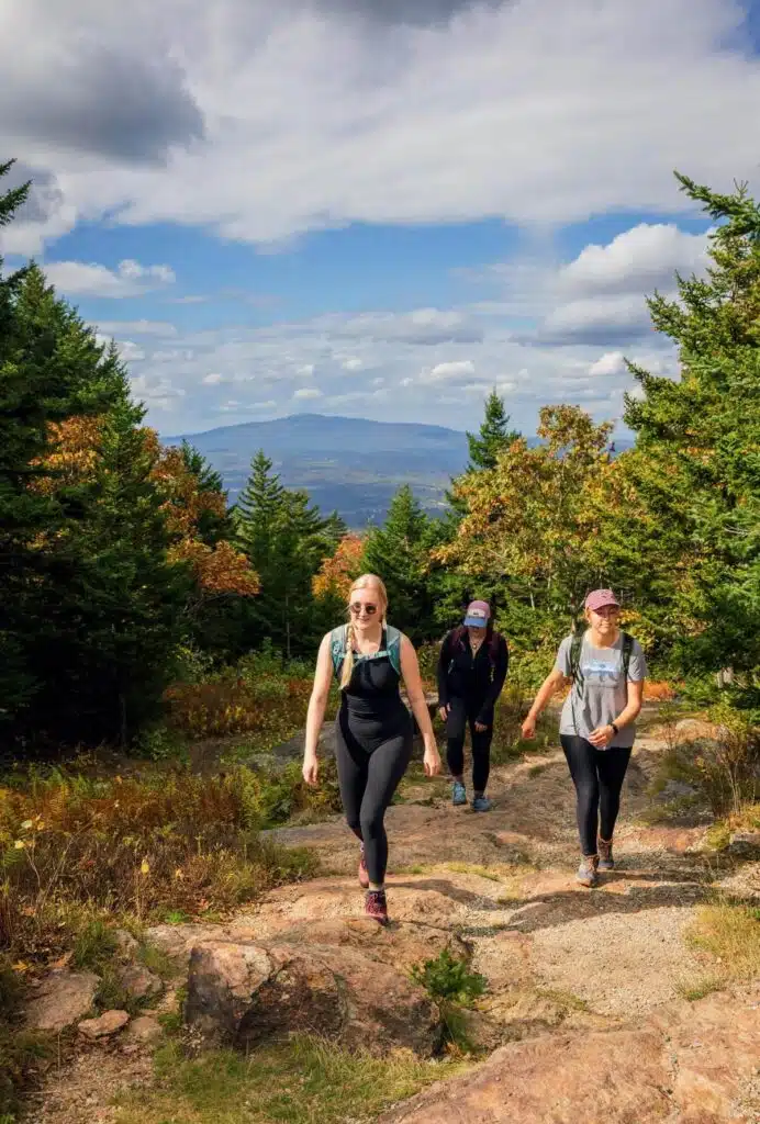 Three women hike up a rocky trail surrounded by trees with mountains in the background under a partly cloudy sky.