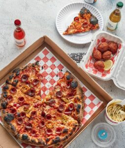 Open pizza box with pepperoni pizza, sliced, with drinks, garlic knots, and a plate with a pizza slice on a table.
