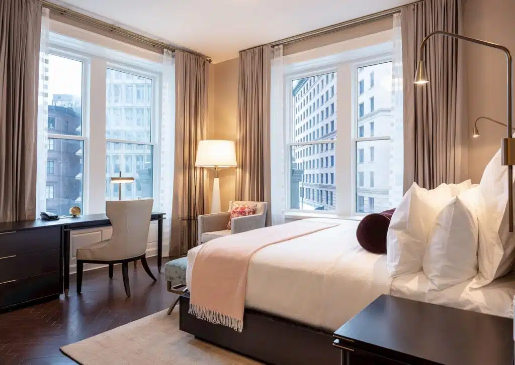 Modern hotel room with large windows, a neatly made bed, desk, chair, armchair, and city views.