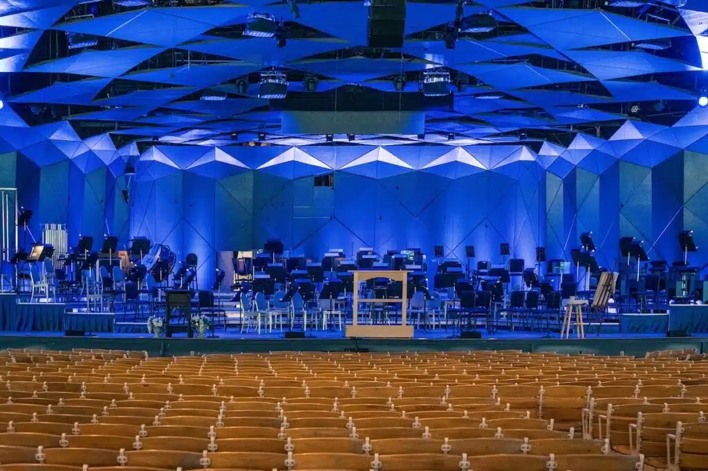 An empty concert hall with a blue-lit stage set for an orchestra, viewed from the audience seats.