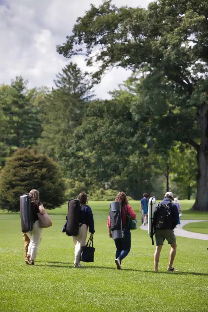 Four people walk across a grassy park area carrying musical instrument cases on their backs on a sunny day.