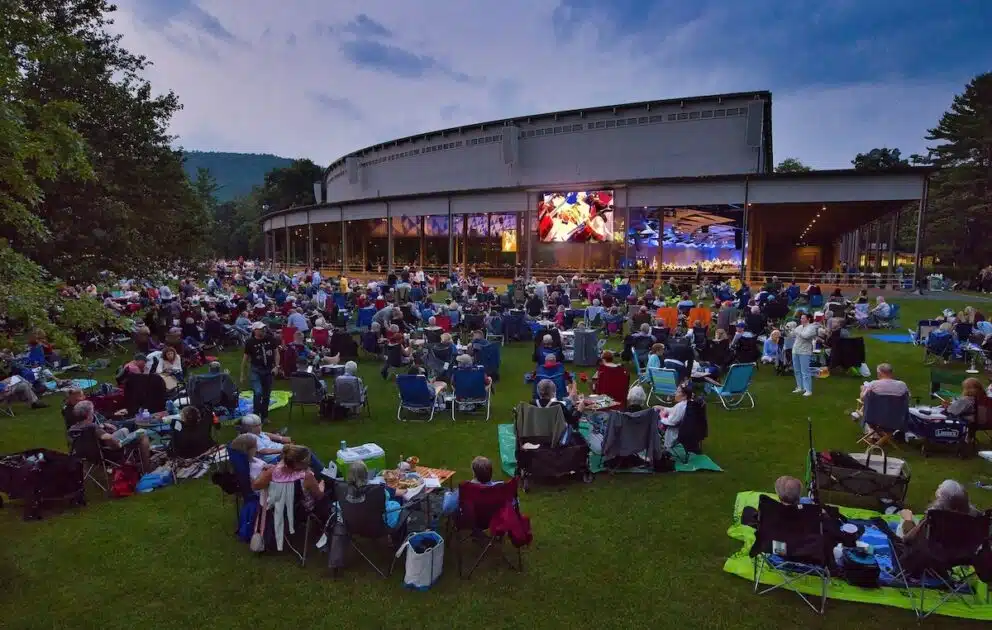 Large crowd sitting on lawn chairs and blankets at an outdoor concert venue during dusk, watching a performance.