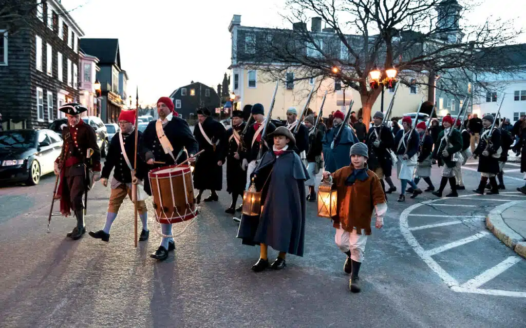People in historical costumes, some holding drums and lanterns, walk in a group on a town street at dusk.