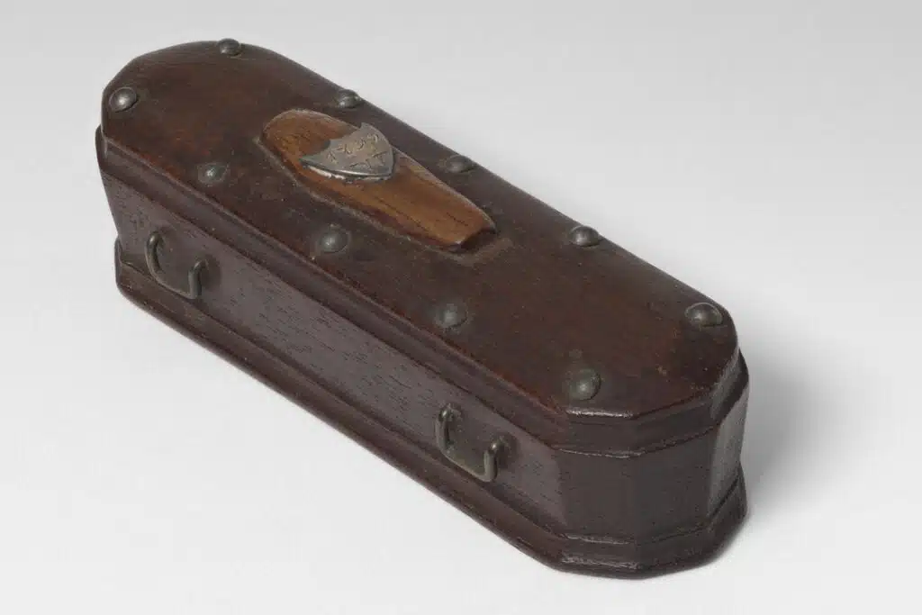Small, dark wooden coffin-shaped box with metal studs, handles, and a decorative shield on the lid.