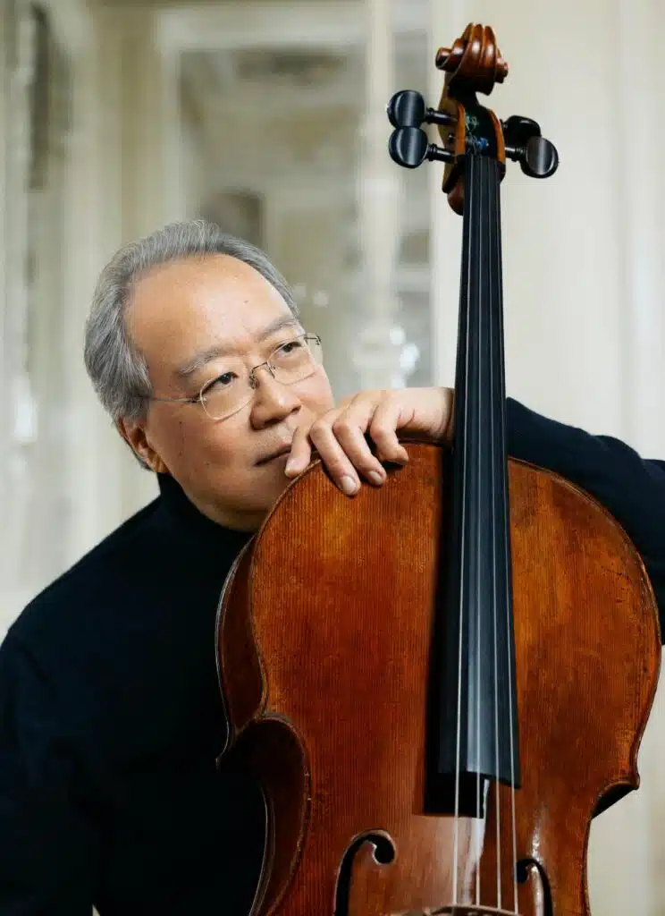 Yo-Yo Ma: An older man with glasses leans on a cello, looking thoughtful, in a softly lit indoor setting. Musician with BSO, Tanglewood, "We the People" weeklong residency.
