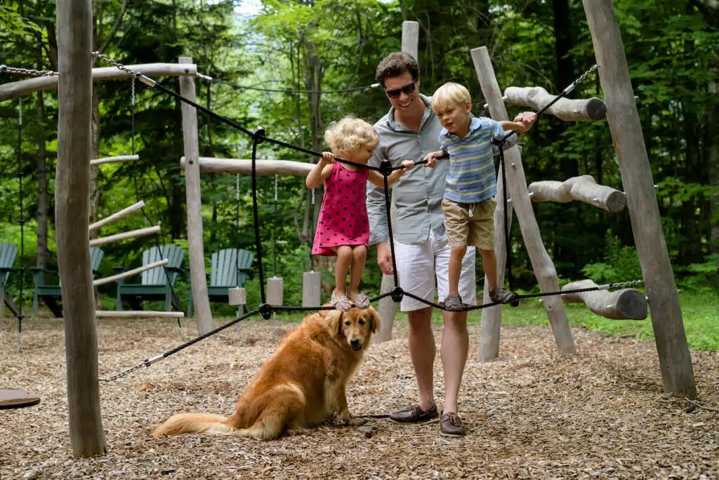 A man helps two young children cross a rope bridge at a playground, with a golden retriever sitting nearby.