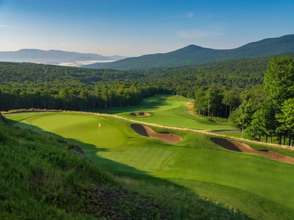 A scenic golf course with lush greens, sand bunkers, and mountains in the background under a clear sky.