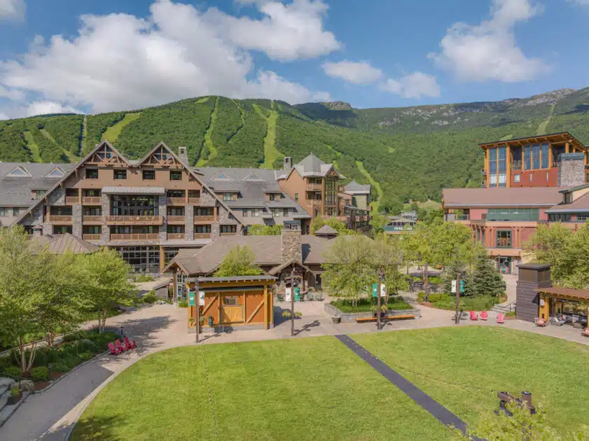 A mountain resort with chalet-style buildings, green lawns, and forested hills under a blue sky with clouds.
