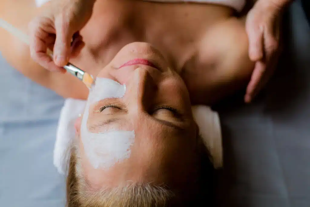 Person lying down with eyes closed, receiving a white facial mask applied with a brush to their forehead.