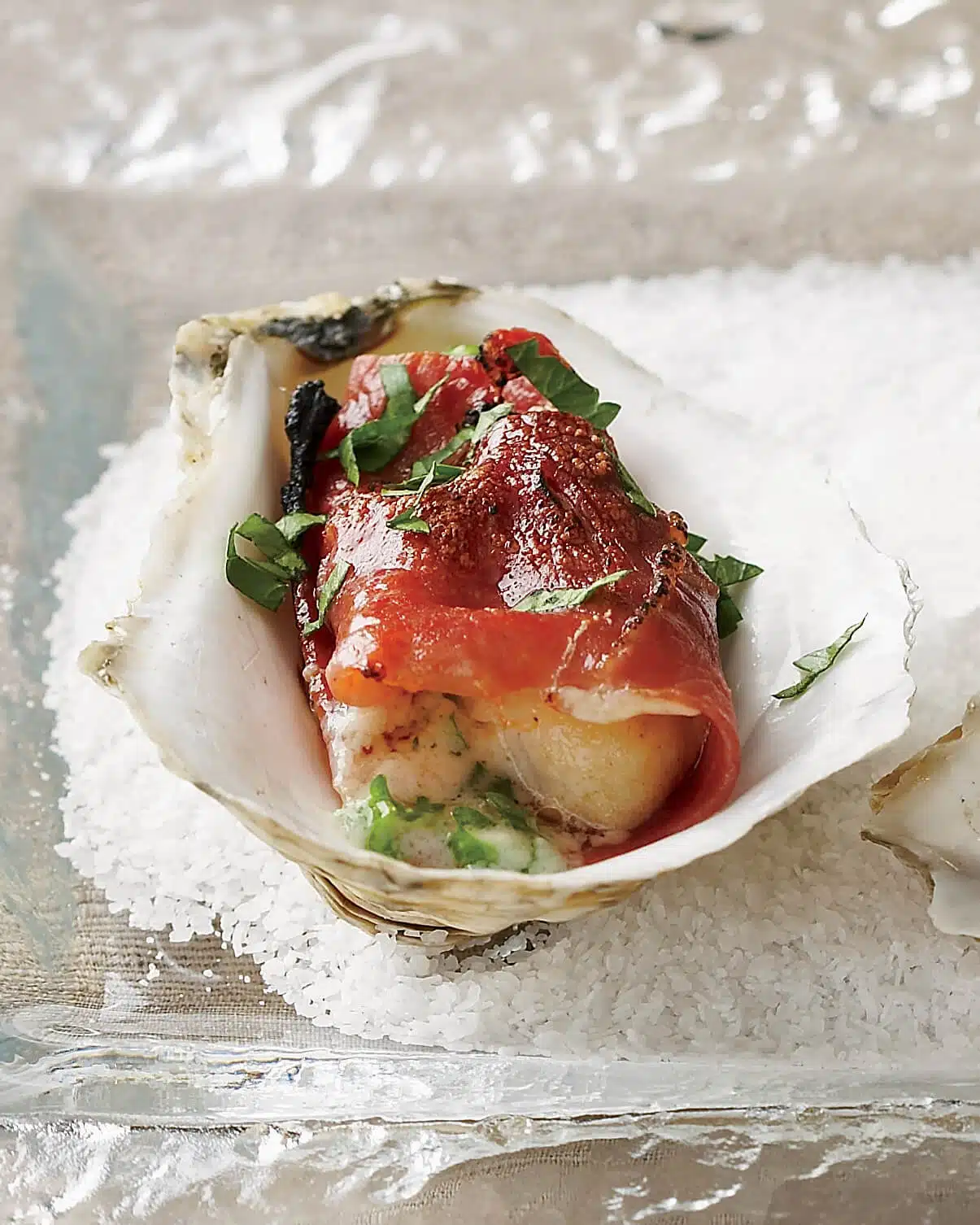 An oyster on a half shell topped with prosciutto and chopped herbs, served on a bed of coarse salt.