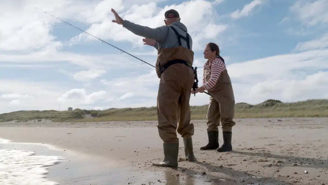 Two people in waders fishing on a sandy beach, one casting a line while the other observes.