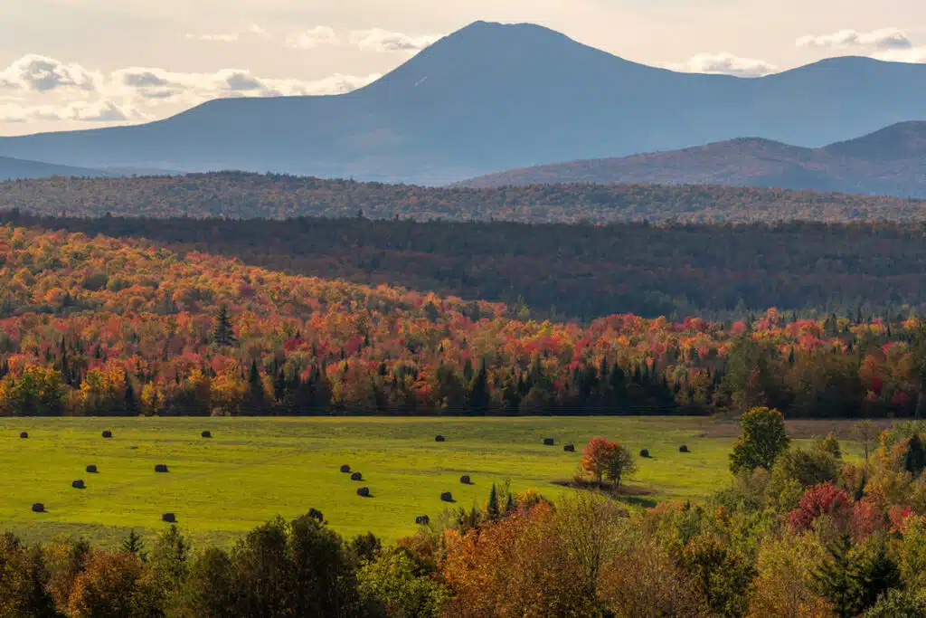 A field with hay bales, colorful autumn trees, and distant mountains under a partly cloudy sky.