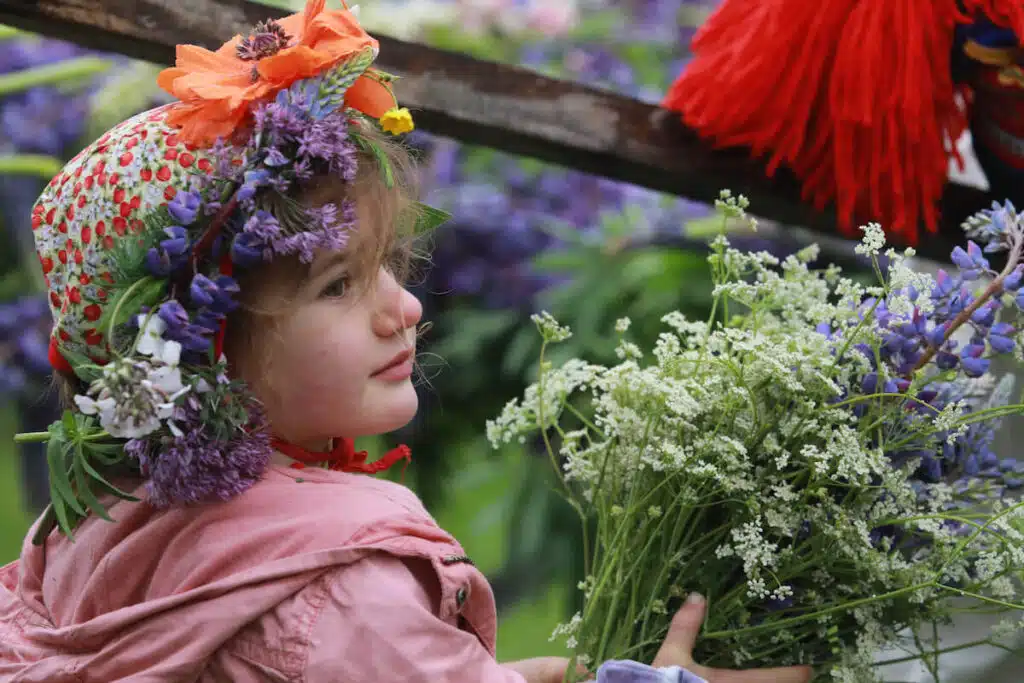 A child in a floral hat holds a bunch of wildflowers, surrounded by colorful blooms outdoors.