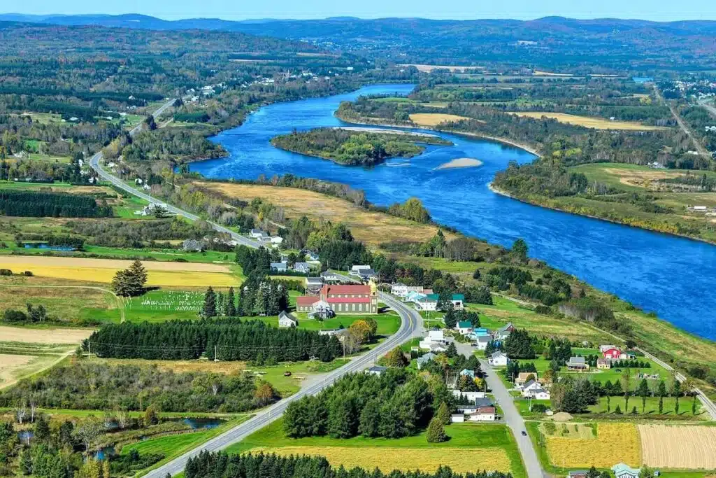 Aerial view of a rural village by a winding river, surrounded by green fields and forested hills.