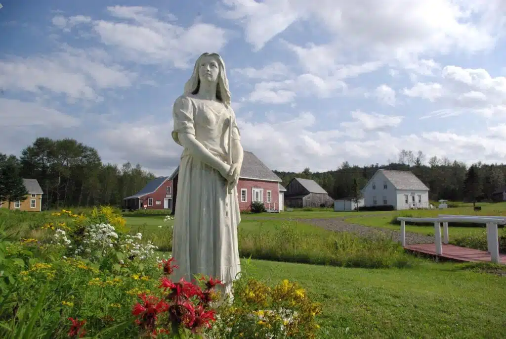 Statue of a woman in a garden with flowers, historic houses, and trees under a partly cloudy sky.