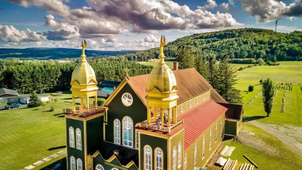 Aerial view of a green church with golden domes amid fields, trees, and distant hills under a blue sky.
