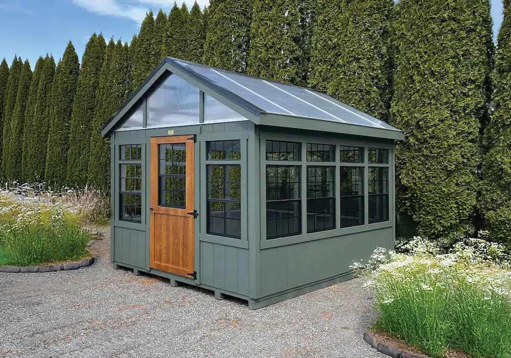 A small green greenhouse with a wooden door, surrounded by tall hedges and wildflowers.